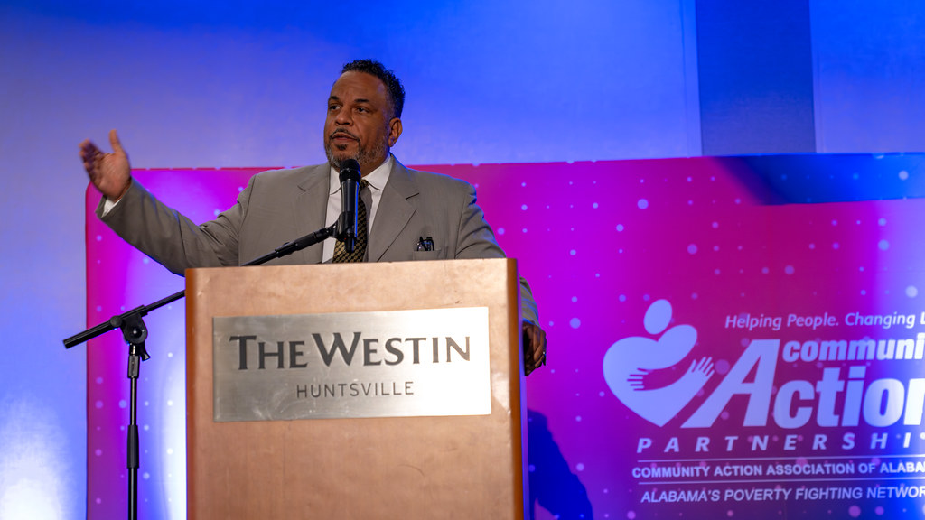 A speaker gesturing from the podium with a Community Action backdrop behind him