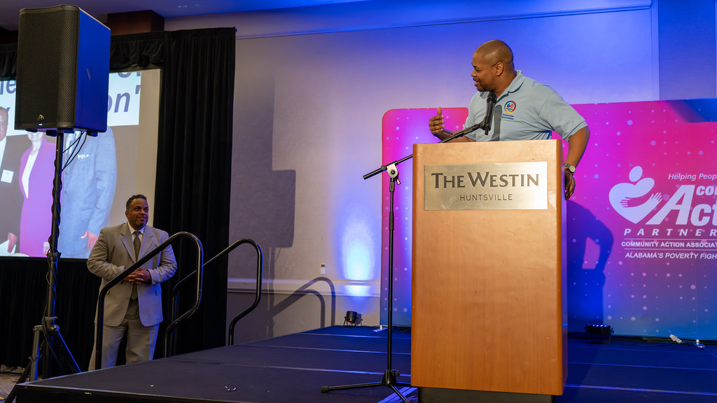 A speaker at a wooden podium on a stage lit in blue with a Community Action backdrop
